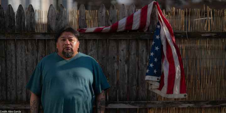 Margarito Casta​​ñon Nava looks into the camera as he stands next to an American flag hanging from a wooden fence behind him on his right.