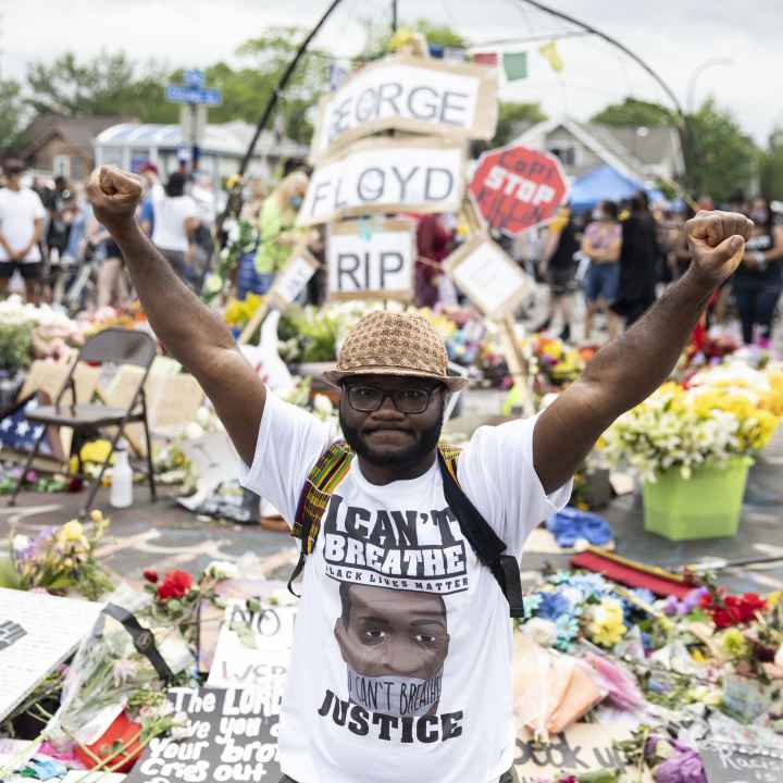 Man wearing "I Can't Breathe" tshirt stands in front of George Floyd memorial with fists raised.