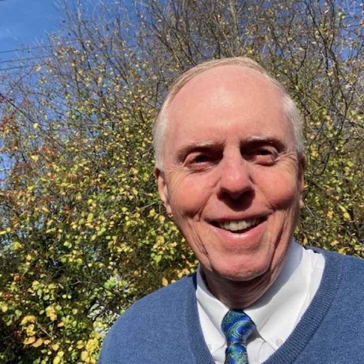 Photo of Bruce wearing a blue sweater and tie in front of a small, green tree.