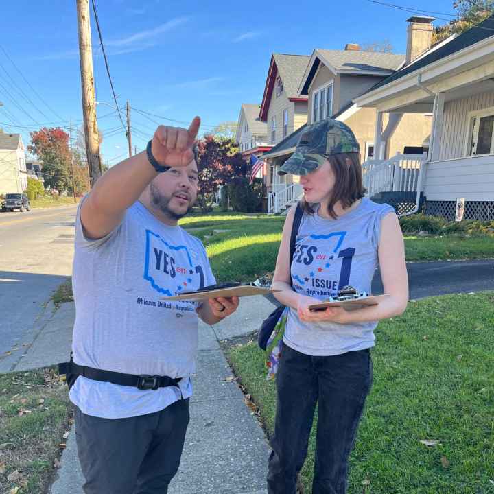 Photo of Julio on left pointing and talking to a woman on the right. Both are wearing gray t-shirts that say "yes on issue 1" with the shape of the state of Ohio behind the text.