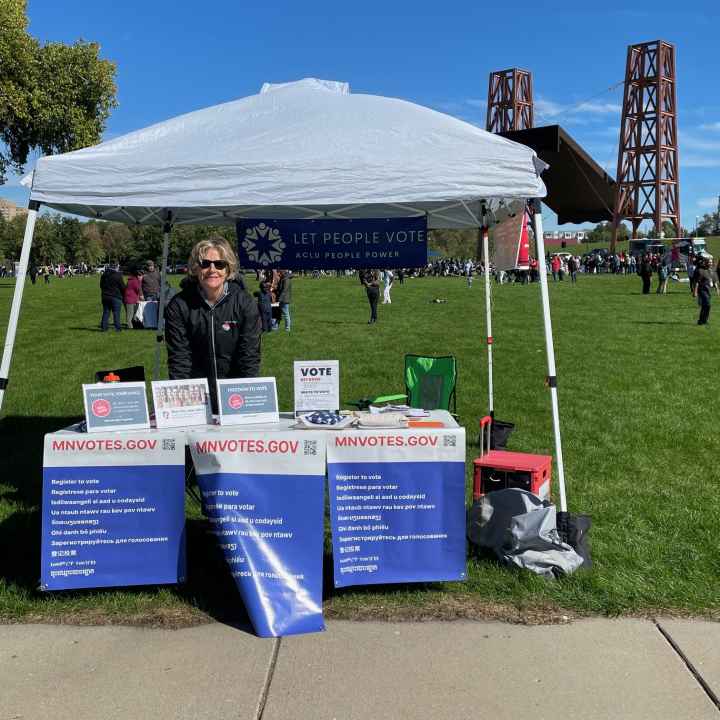 Photo of an individual with short blond hair, wearing sunglasses, standing under a white tent. In front of the person is a table with signs attached to it. The signs say, "MNvotes.gov." 