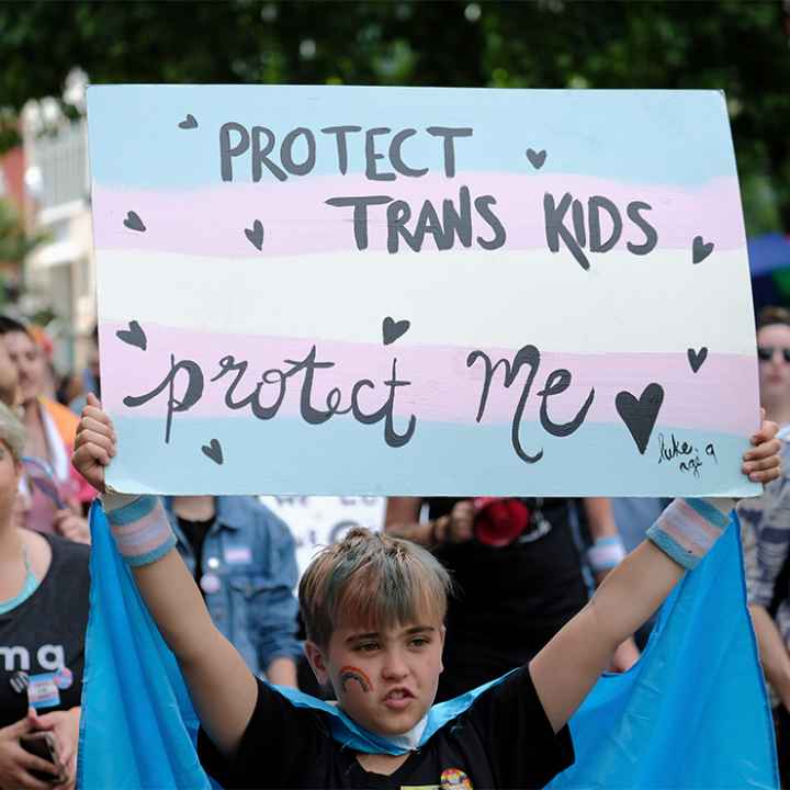 A young child holds up a sign reading "PROTECT TRANS KIDS, PROTECT ME - luke, age 9" at a Trans Pride March.